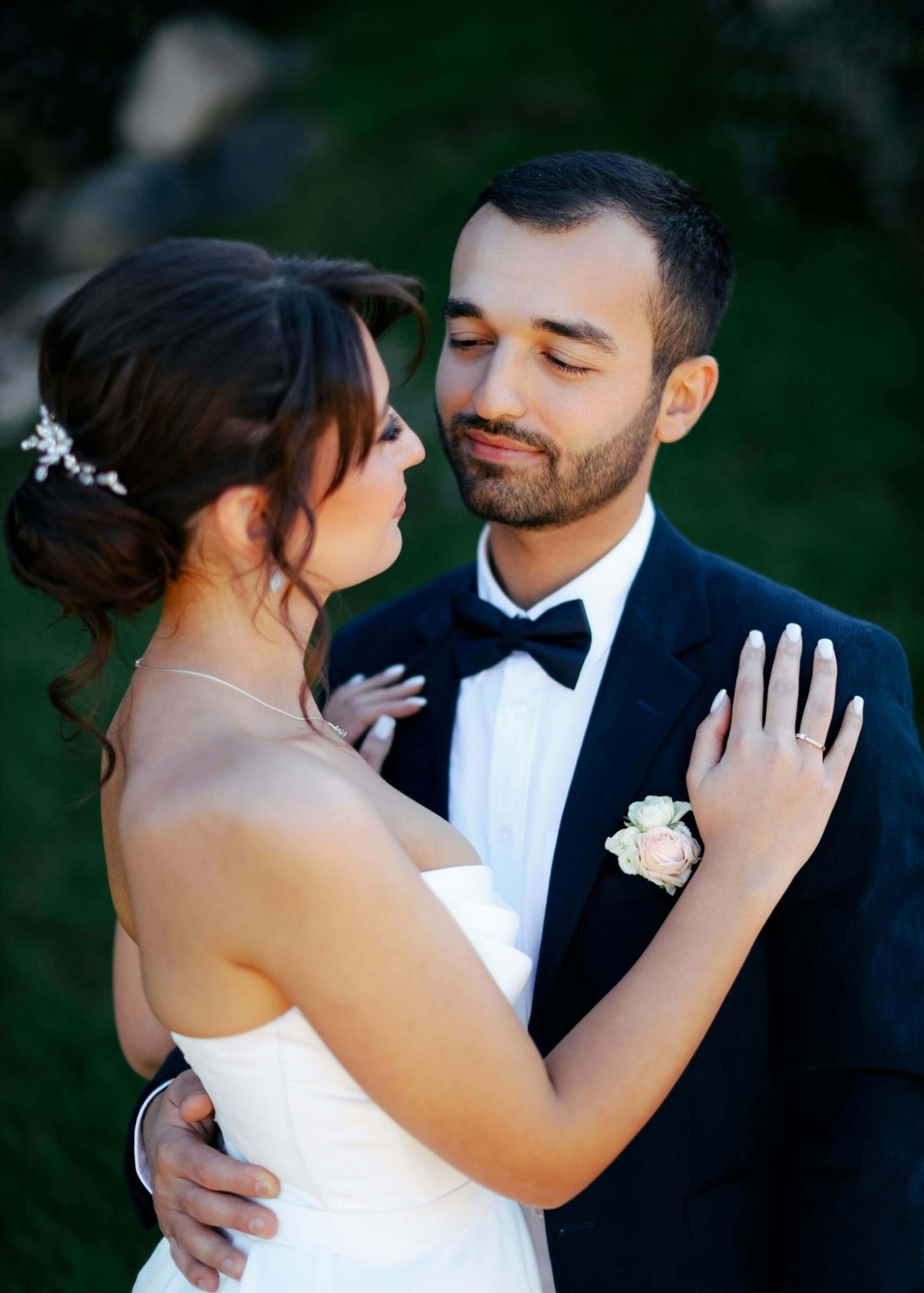Outdoor Embrace Couple in wedding attire embracing outdoors against dark greenery.
