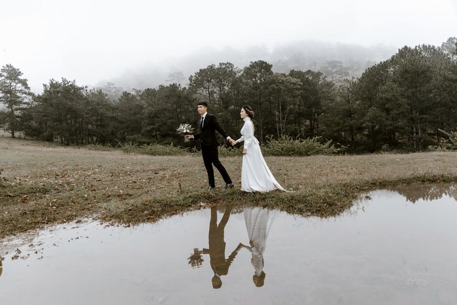 Couple in wedding attire walking beside water with their reflections visible.