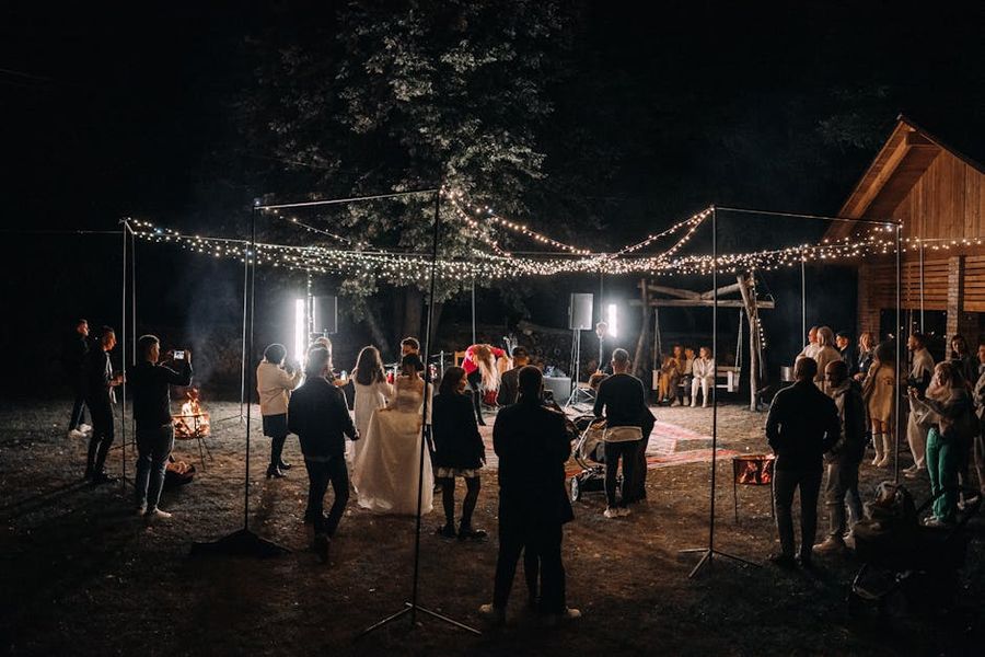 Evening Gathering People gathered beneath string lights around small fires beside a wooden building at night.
