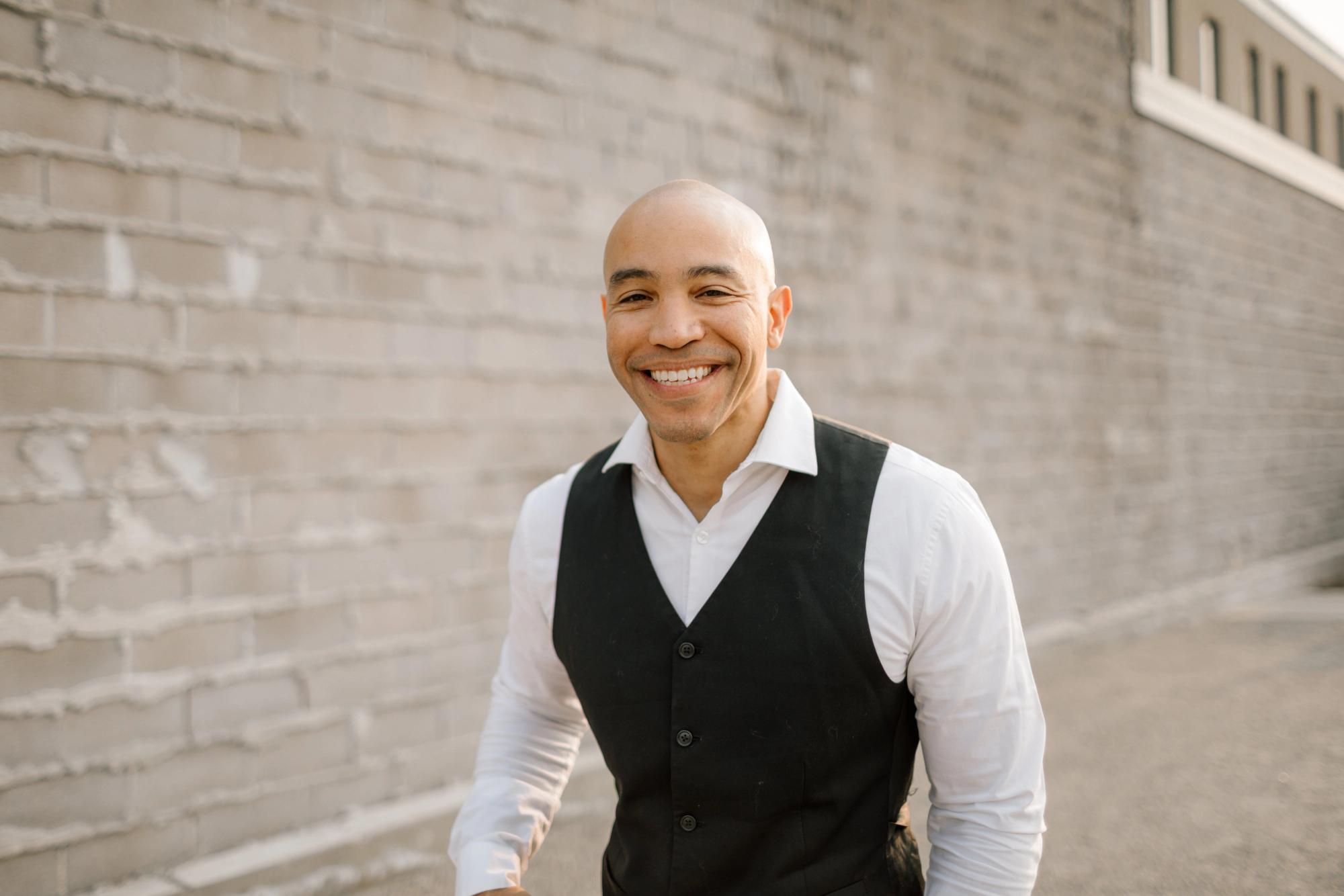 Vest Portrait Duplicate Smiling man in a black vest and white shirt standing beside a pale brick wall.