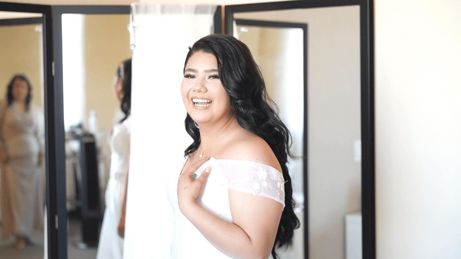 Smiling person in an off-shoulder white dress standing near mirrors.