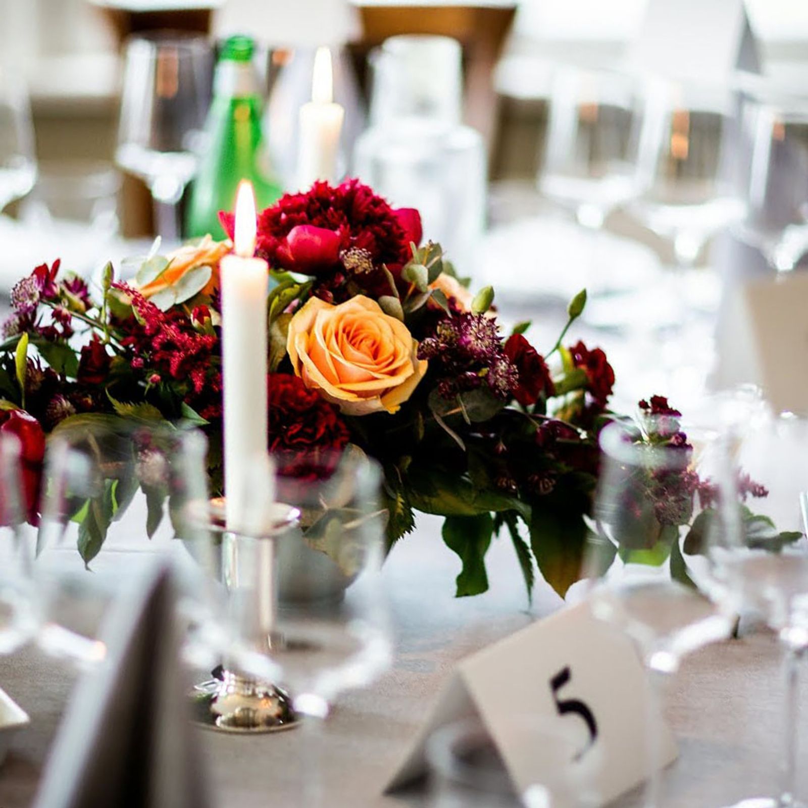 Reception table with red and orange flowers, a candle, glassware, green bottle, and table number five.