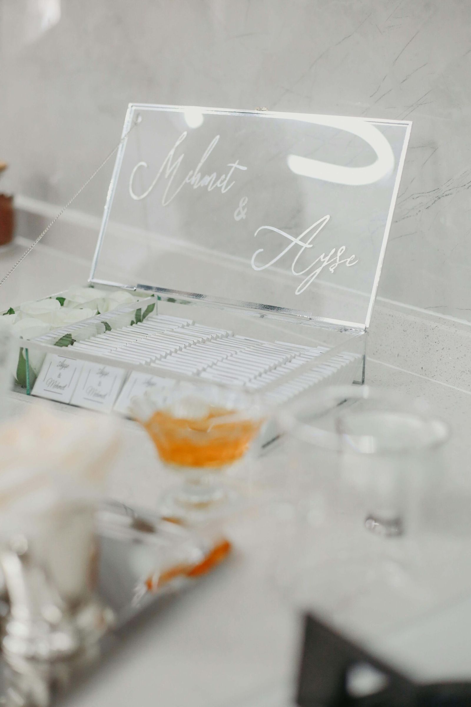 Clear acrylic card box with a script inscription on a table beside small glass containers.