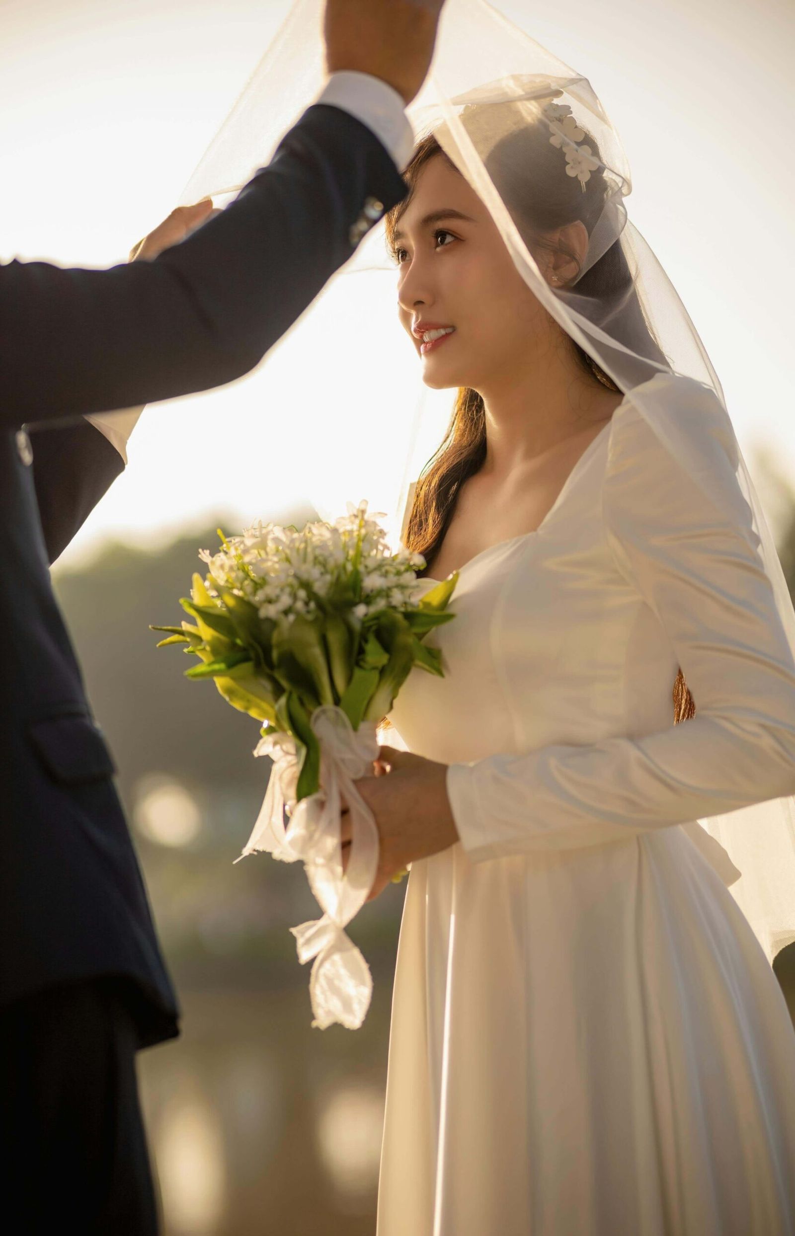 Person in a dark suit lifting a sheer veil from a person holding a green bouquet.