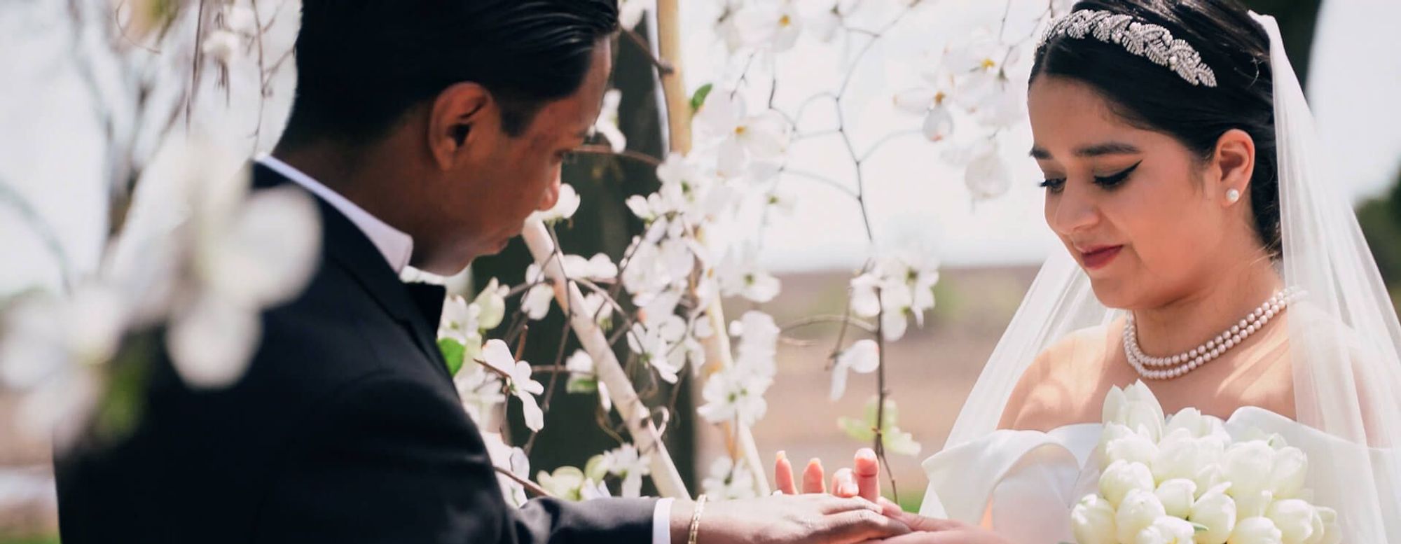 Close crop of one person placing a ring on another person's hand beside white flowers.