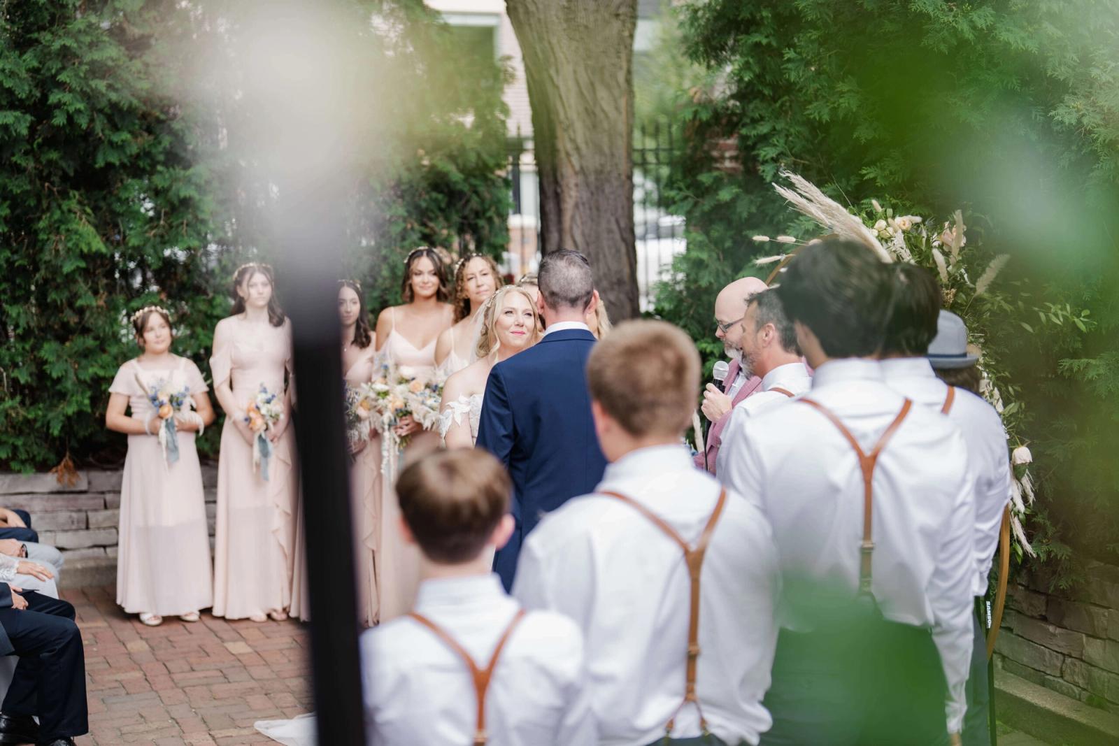 Outdoor ceremony group gathered beneath trees around a couple and officiant.