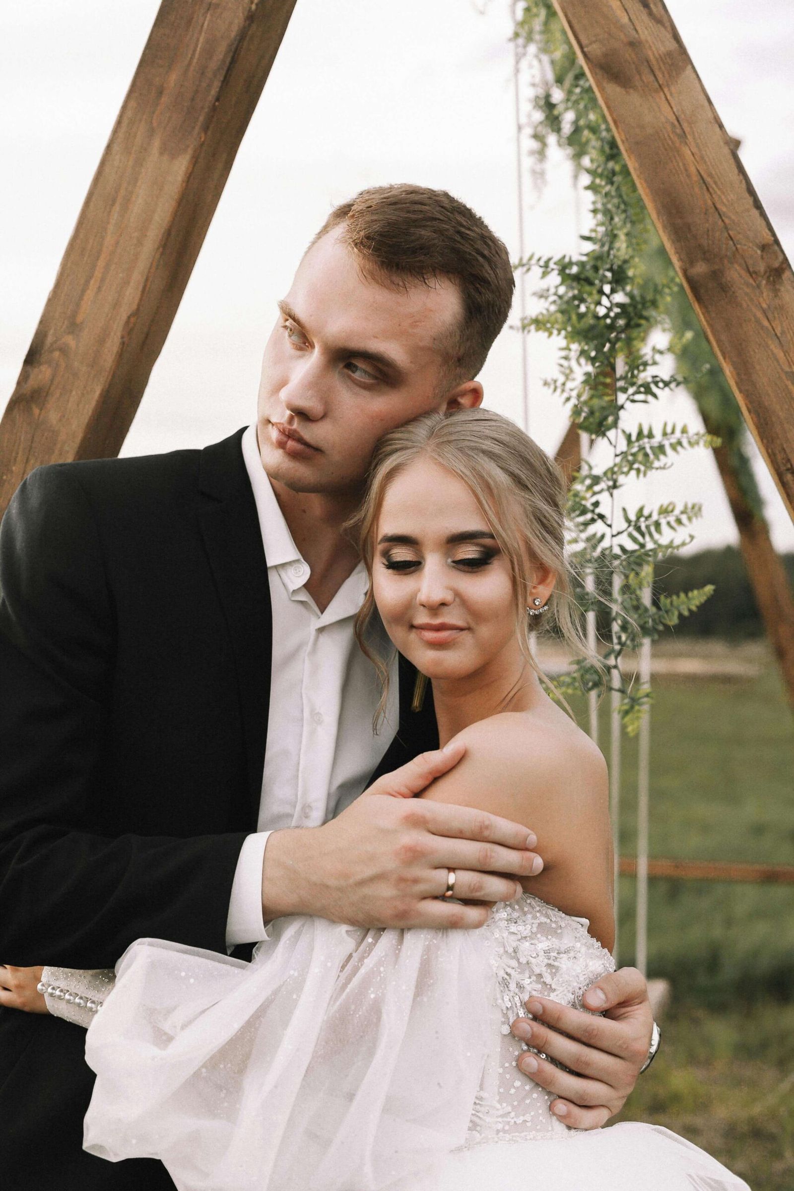 Couple in wedding attire embracing outdoors beside a wooden structure.