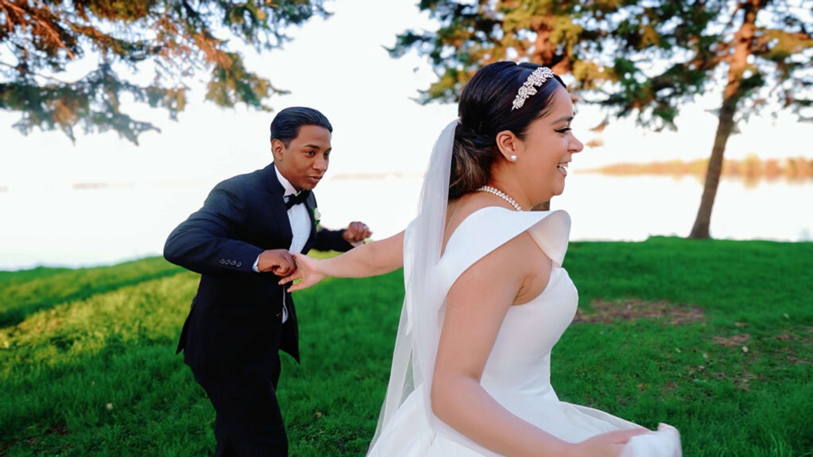 Person in a dark suit playfully running behind a person in a white dress on grass beside water.