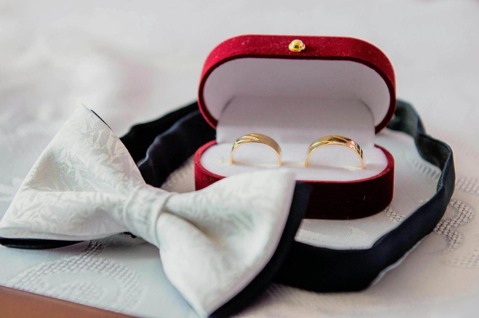 Open red ring box holding two gold bands beside a black bow tie.
