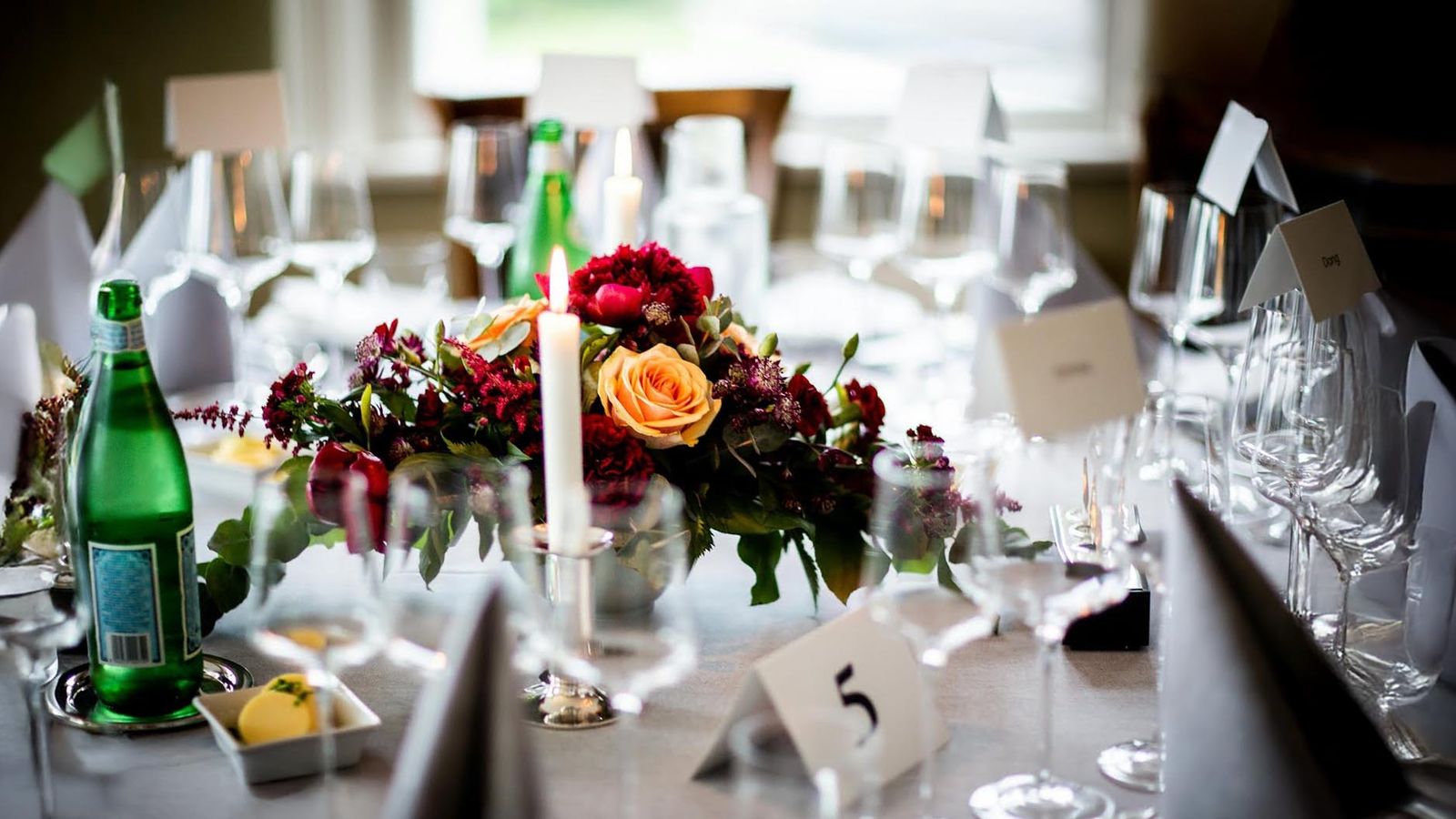 Long reception table with burgundy flowers, candles, glassware, and place cards.