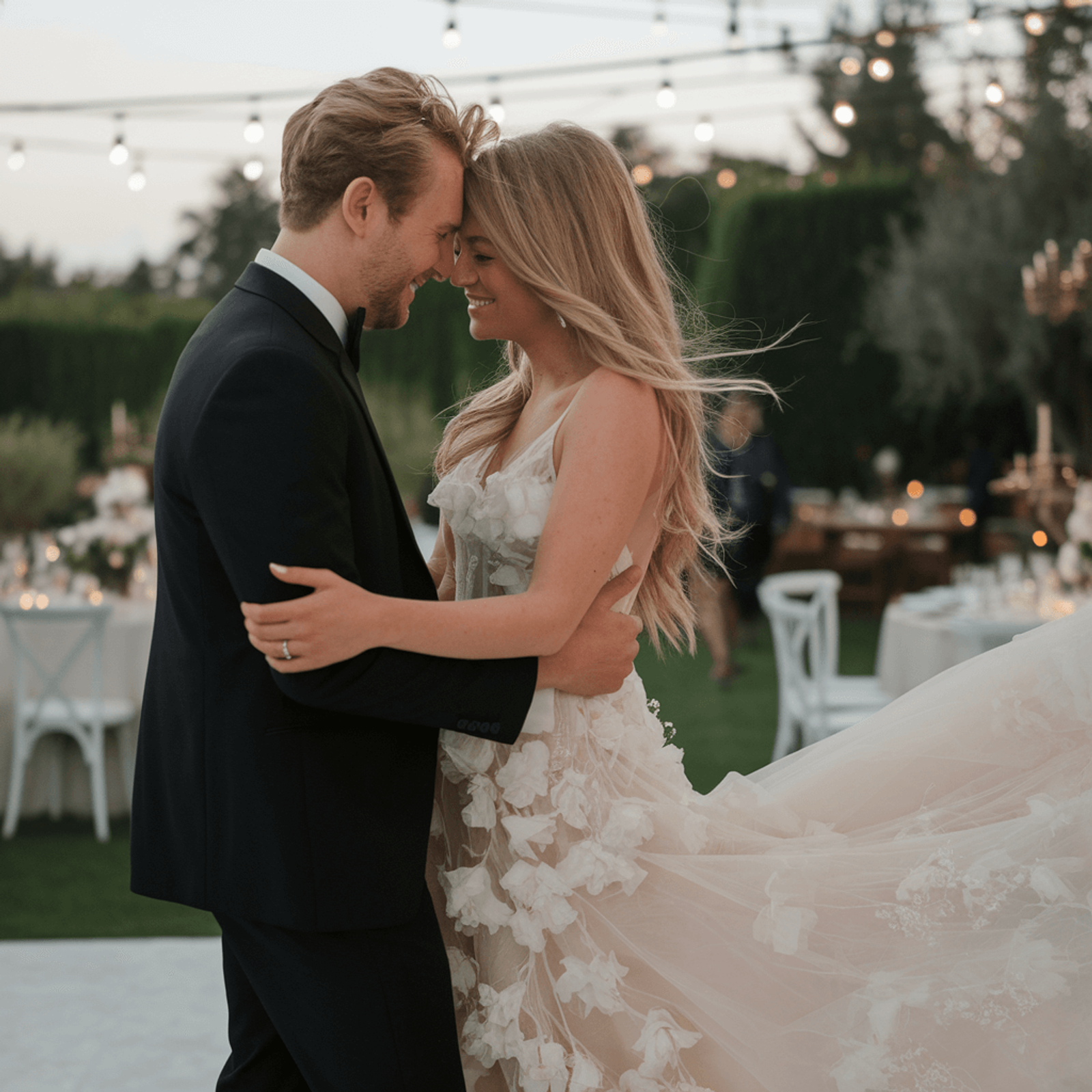 Couple in wedding attire touching foreheads beside an outdoor reception table.
