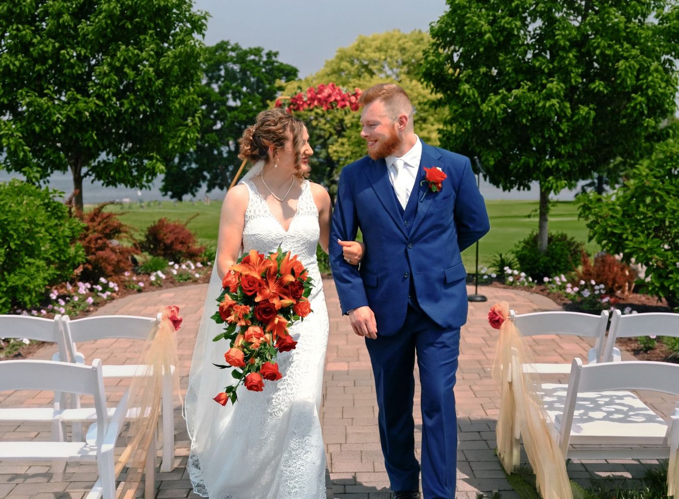 Two formally dressed people walking between white chairs, one carrying orange flowers.