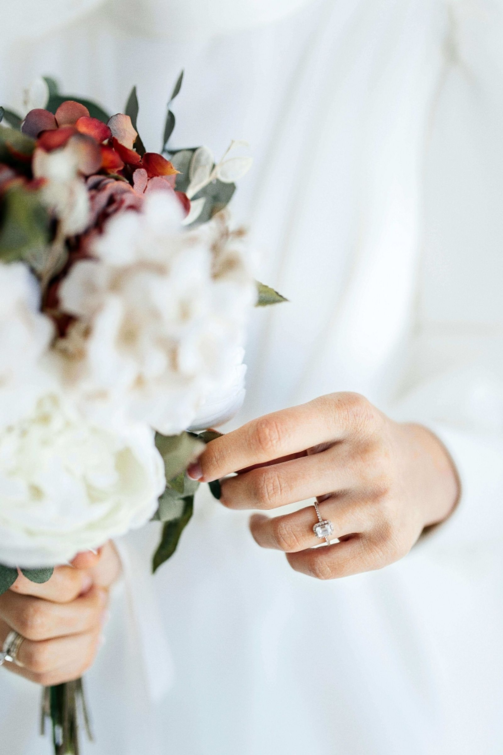 Close-up of a hand with a rectangular gemstone ring holding a white and burgundy bouquet.