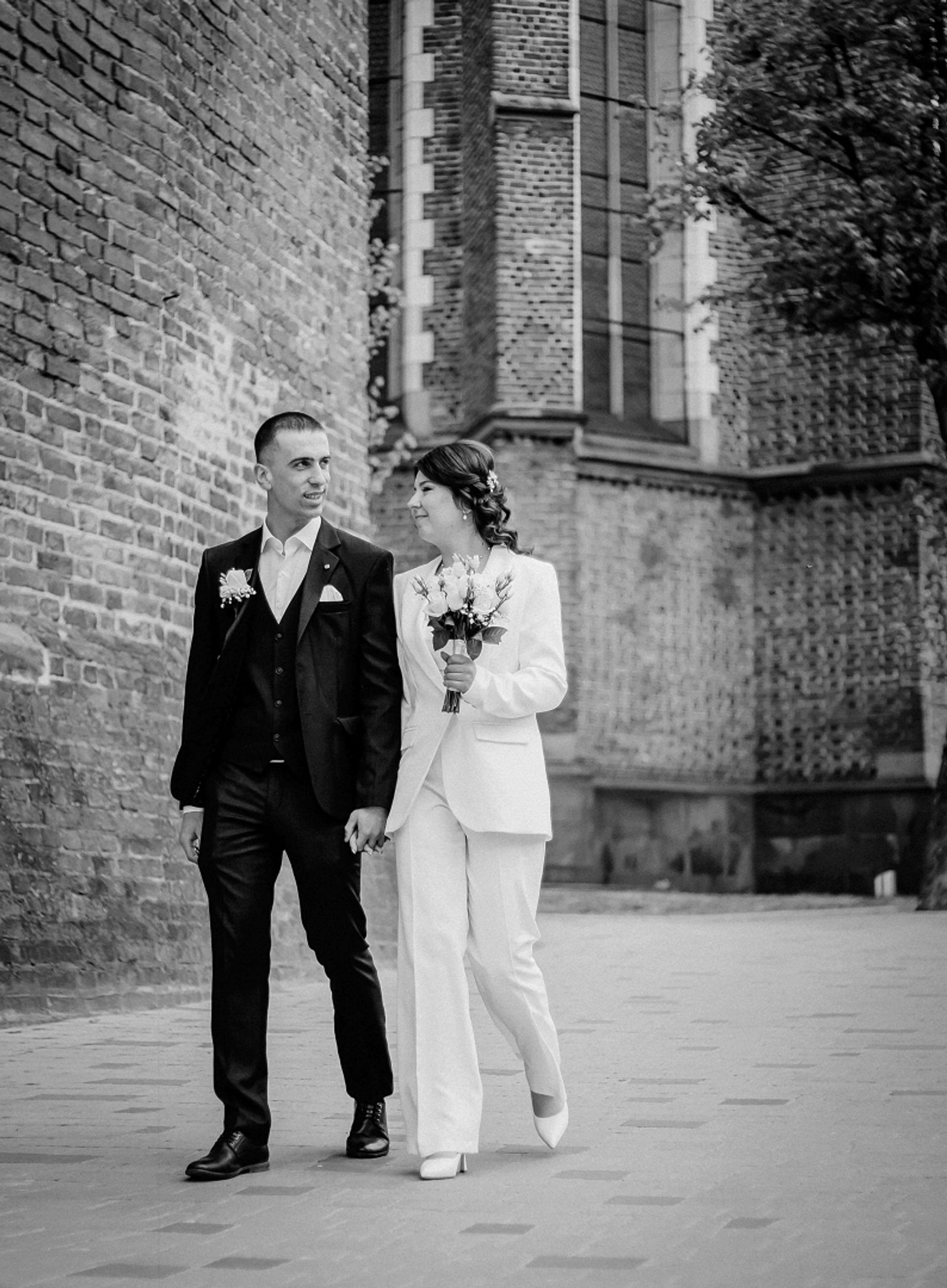Black-and-white image of a couple in wedding attire walking beside a tall brick building.