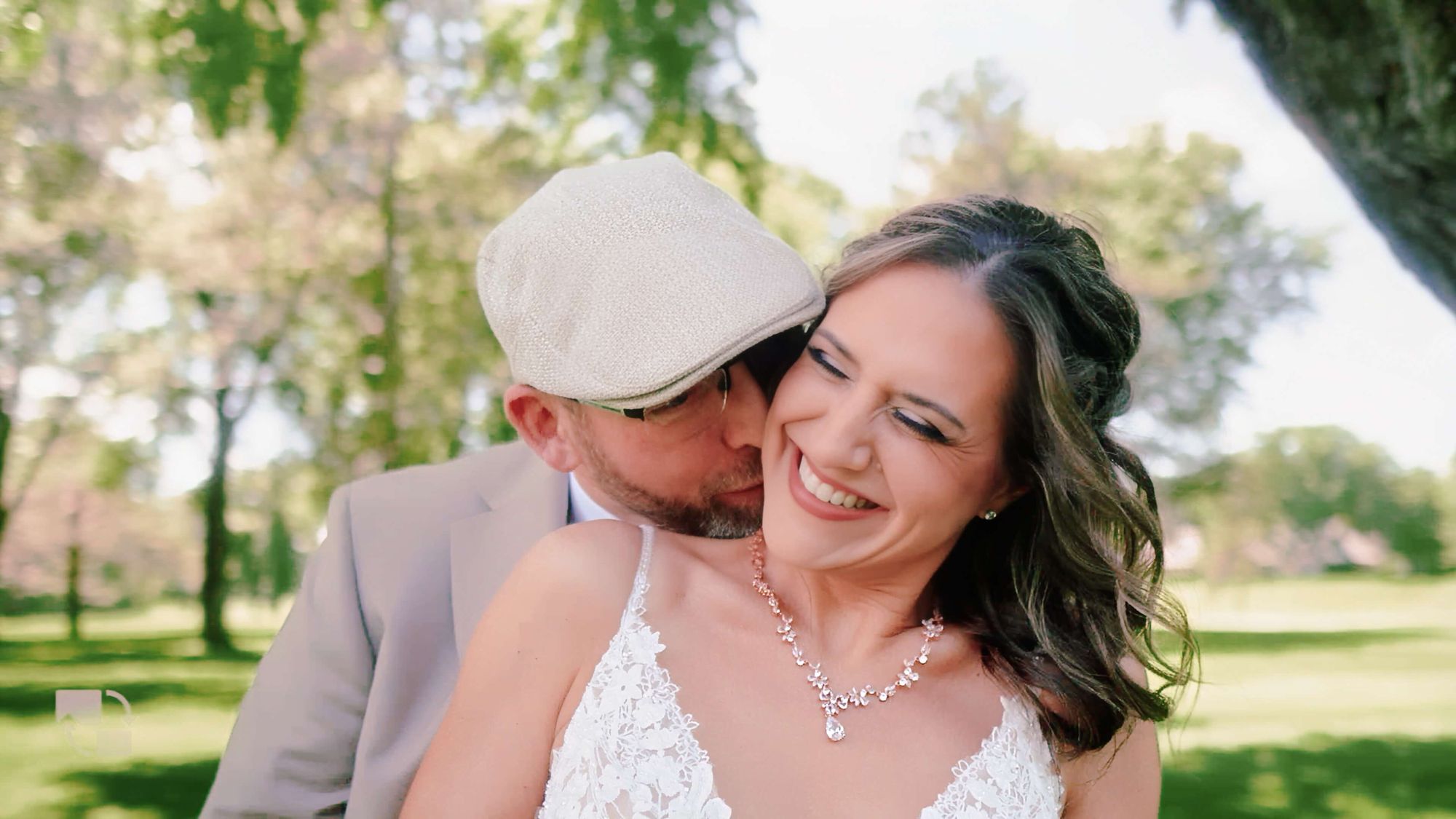 Cheek Kiss Person in a pale cap kissing another person's cheek outdoors beneath trees.