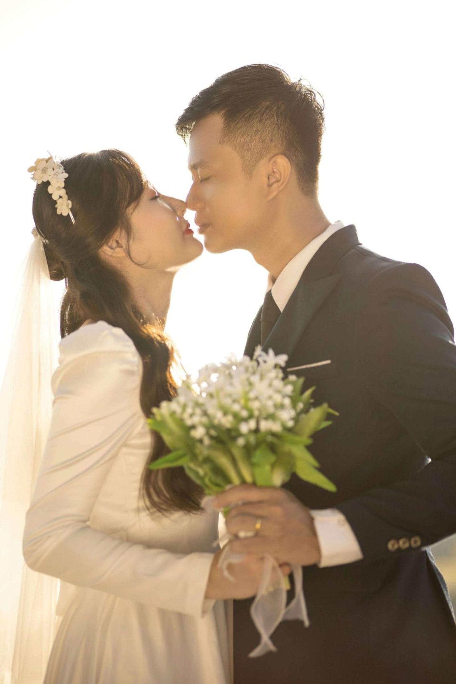 Backlit couple in wedding attire touching foreheads while one holds a bouquet.