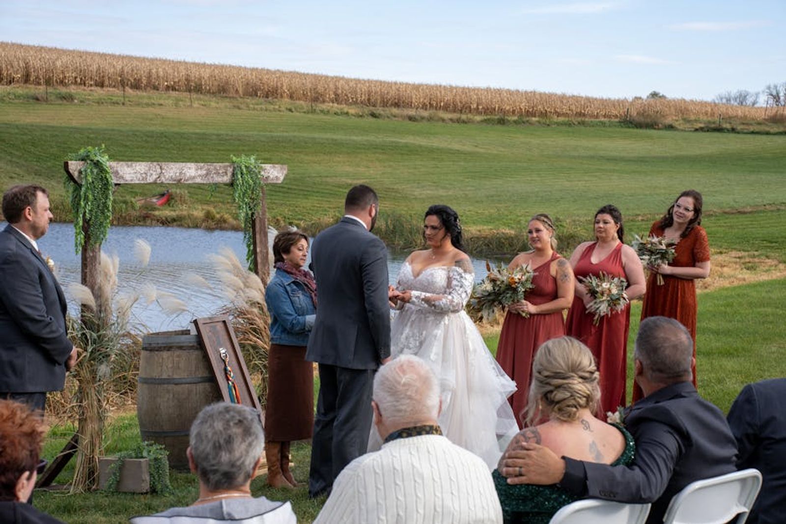 Outdoor ceremony beside a cornfield with guests seated around a floral arch.