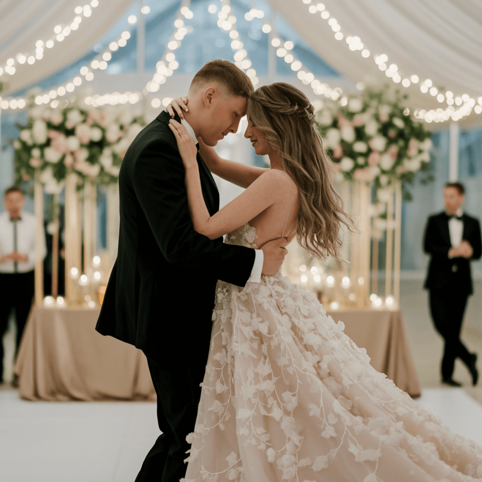 Couple in wedding attire touching foreheads beneath string lights during an indoor dance.