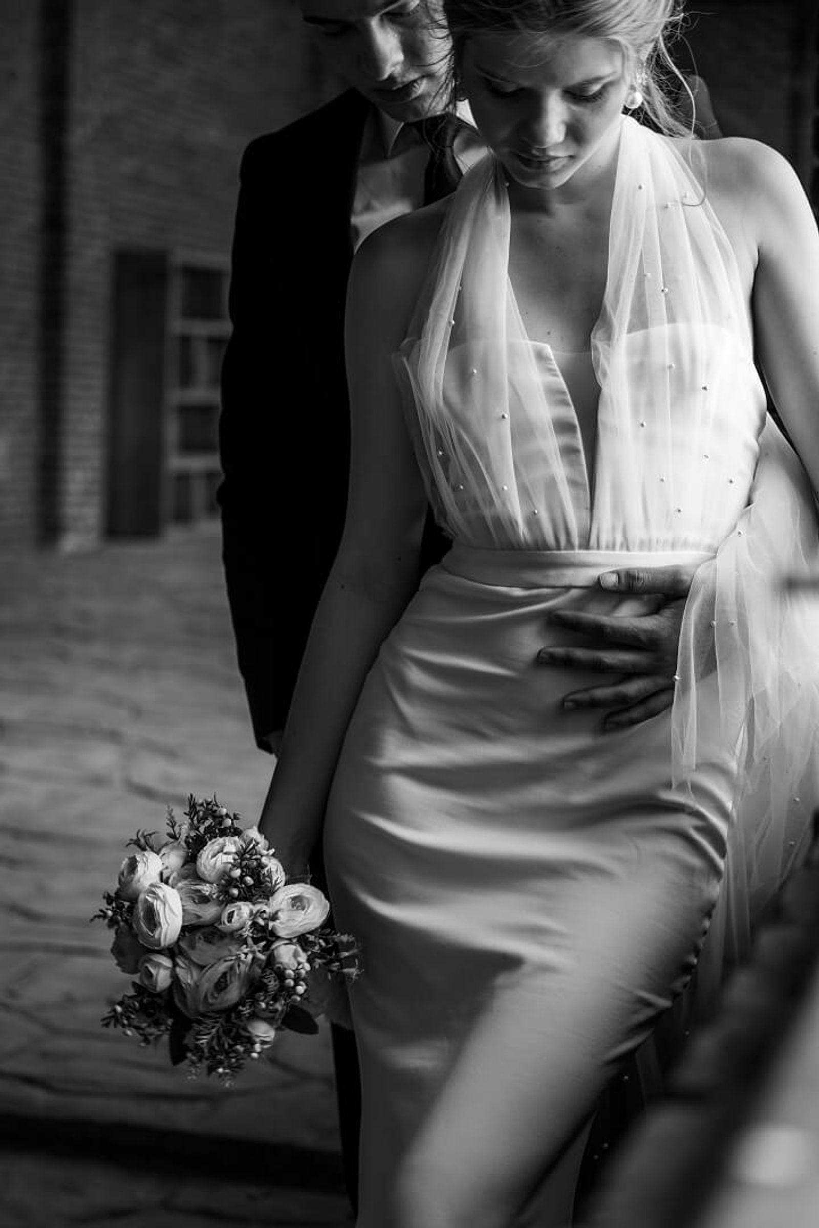 Black-and-white image of a person in a white dress holding a bouquet while another person stands behind with a hand at the waist.