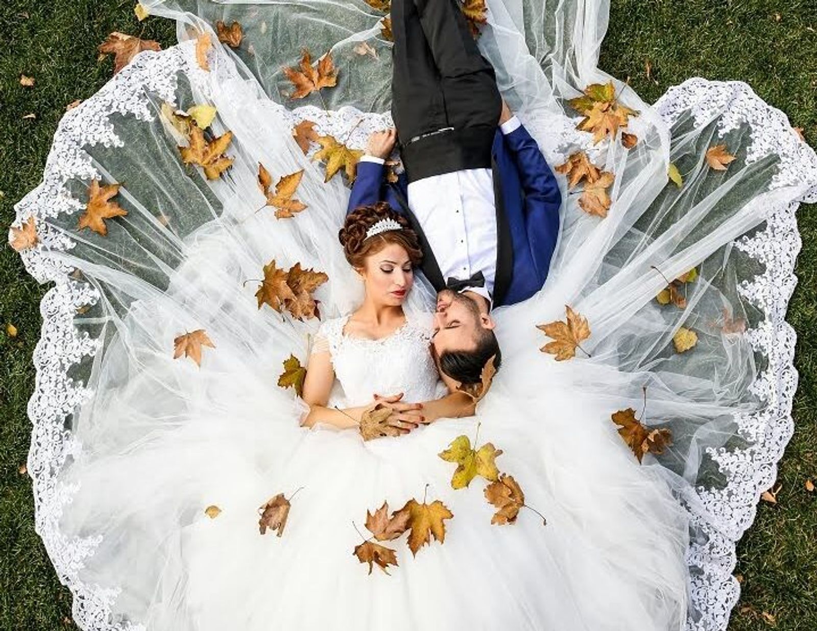 Overhead view of a couple lying on grass, surrounded by a spread white dress and autumn leaves.