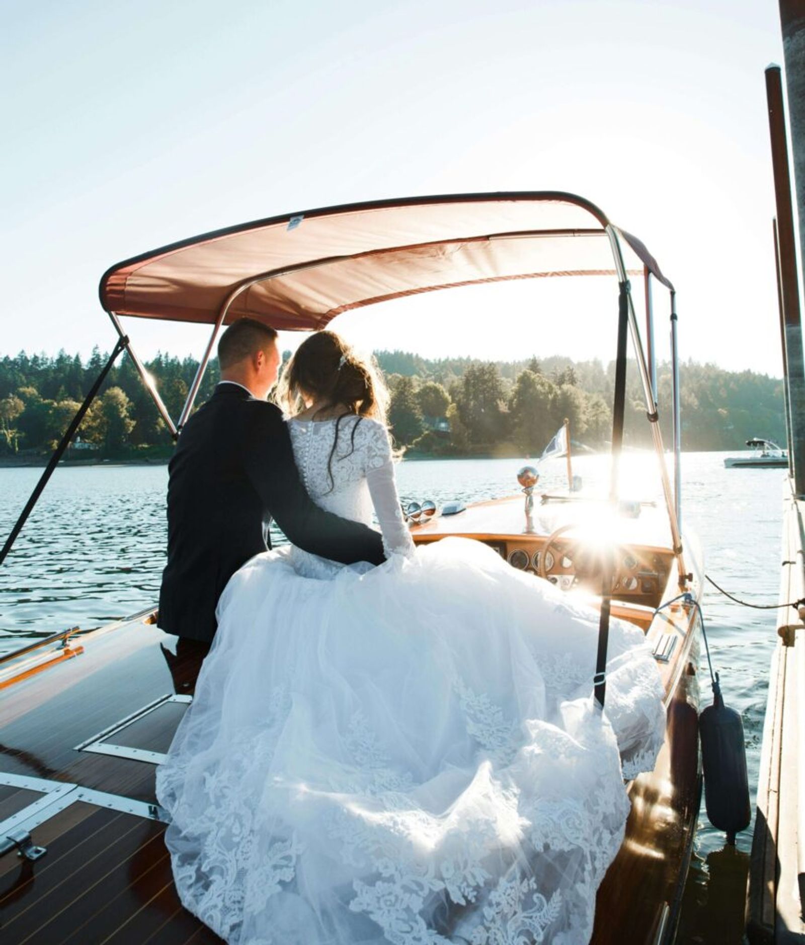 Couple in wedding attire seated together at the rear of a small boat on a lake.