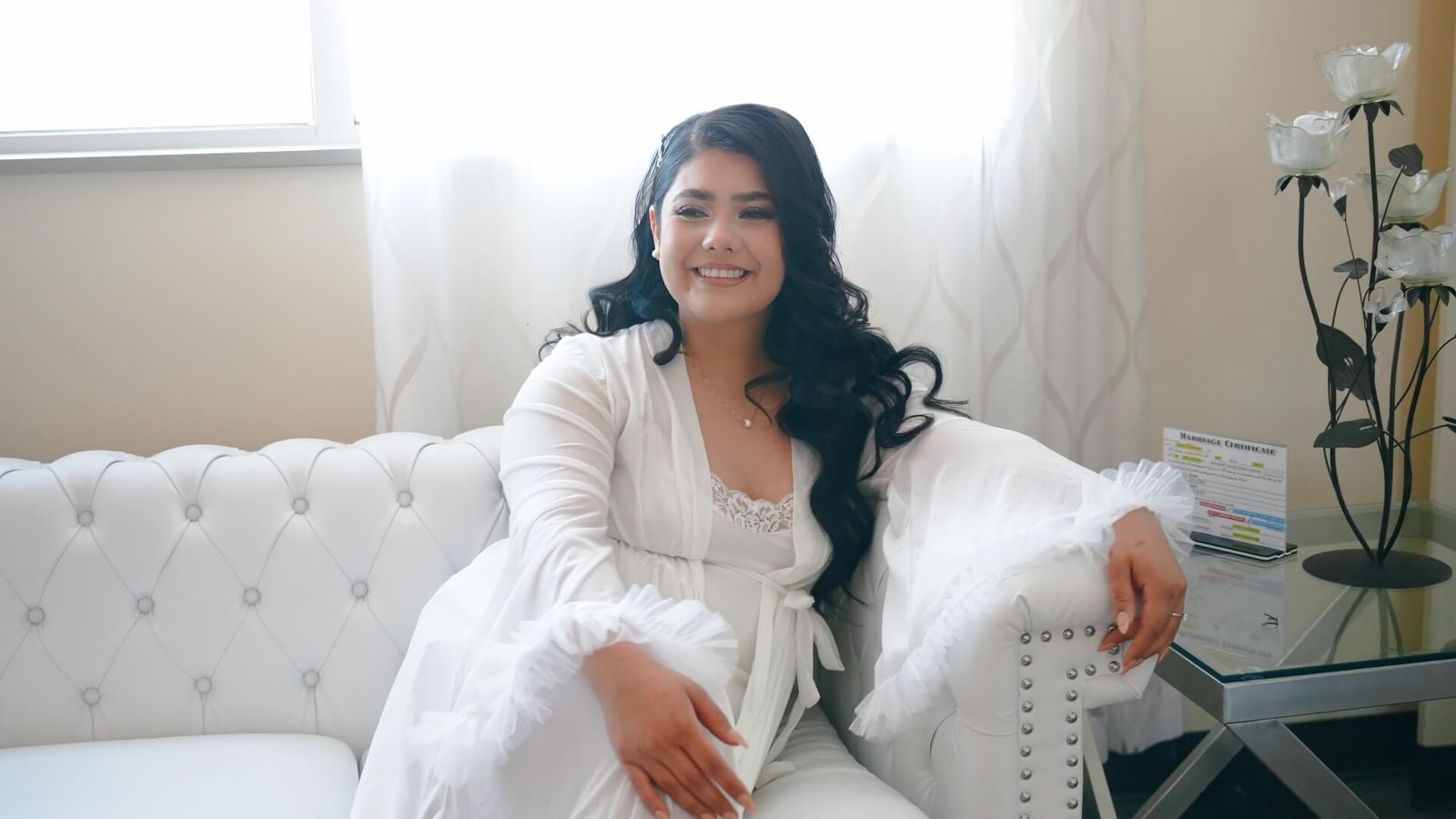 Smiling person in a white robe seated on a white tufted sofa in a bright room.