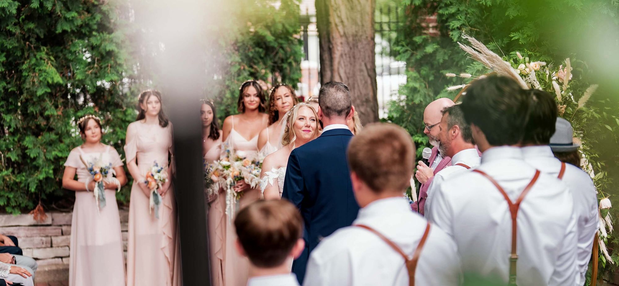 Rear view of seated and standing people watching an outdoor ceremony beneath trees.