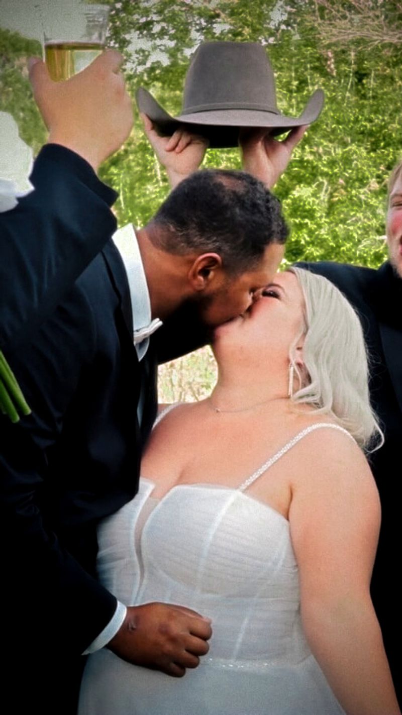 Two formally dressed people kissing outdoors with a person in a cowboy hat behind.