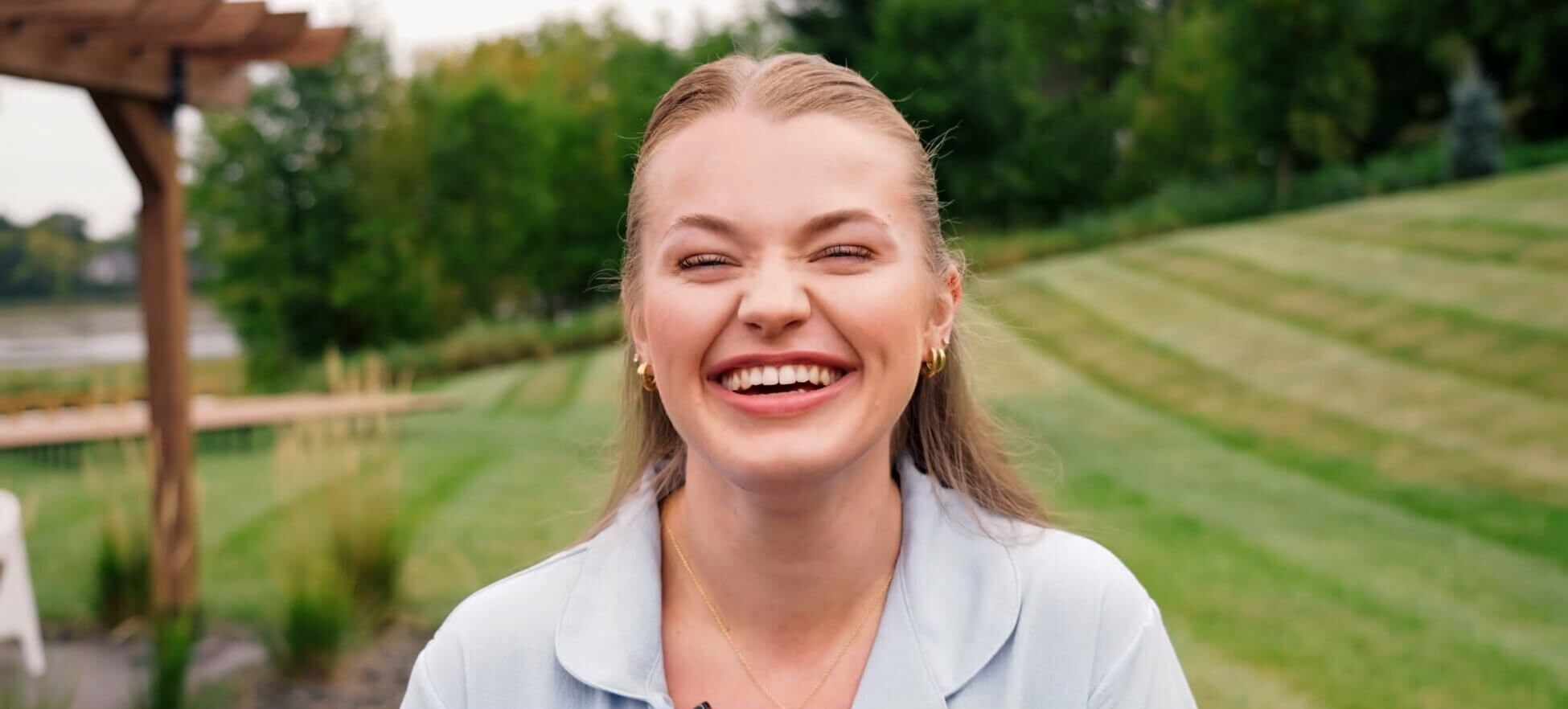 Laughing Portrait Woman in a light jacket laughing toward the camera with a grassy slope behind.