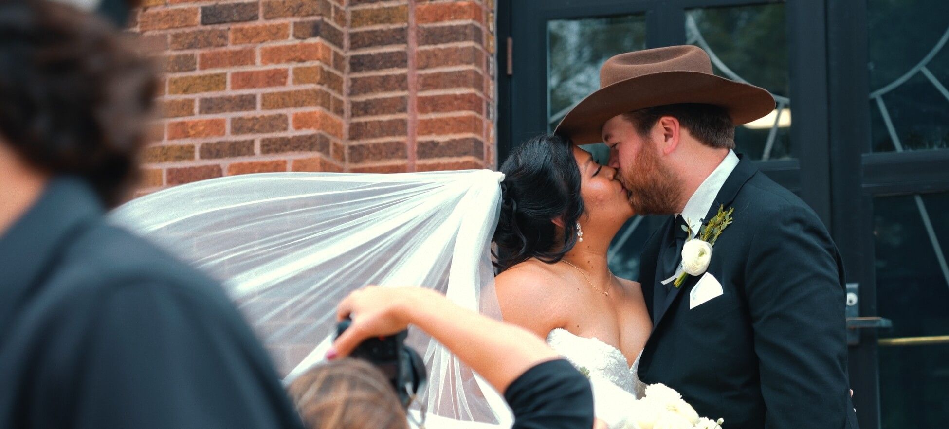 Car Side Kiss Two formally dressed people kissing beside a dark car and brick wall.