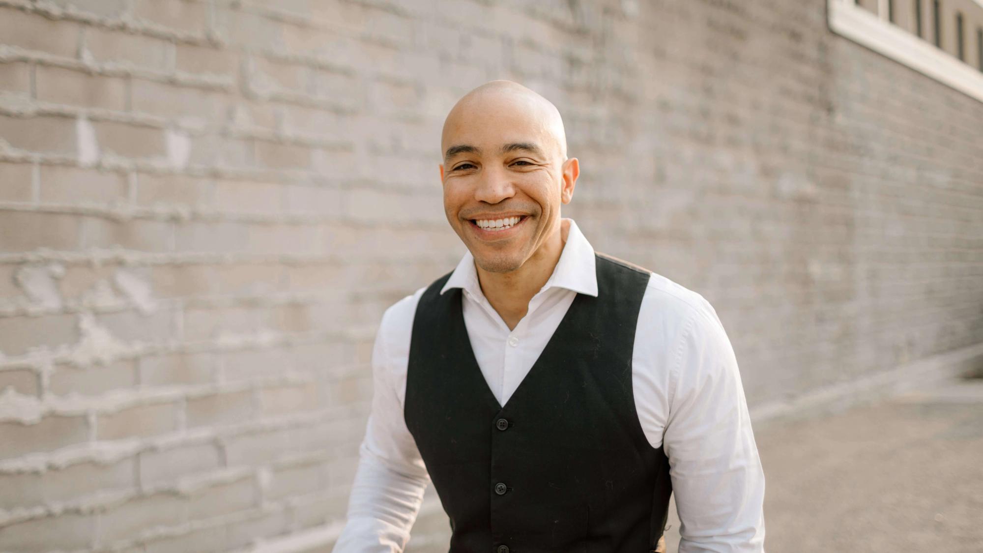 Vest Portrait Third Smiling man in a black vest and white shirt standing beside a pale brick wall.