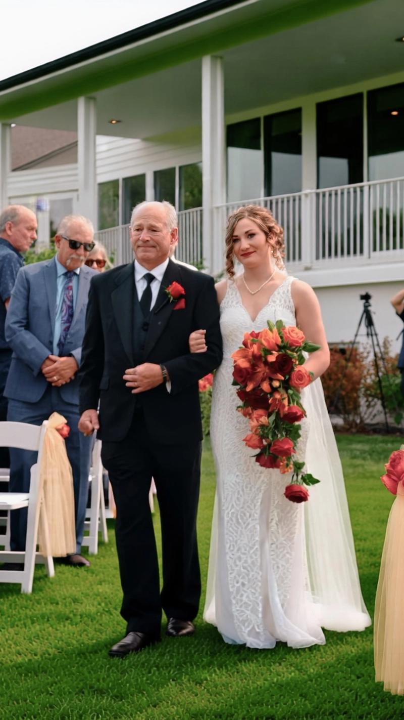 Older man in a dark suit walking arm in arm with a person in a white dress between rows of chairs.