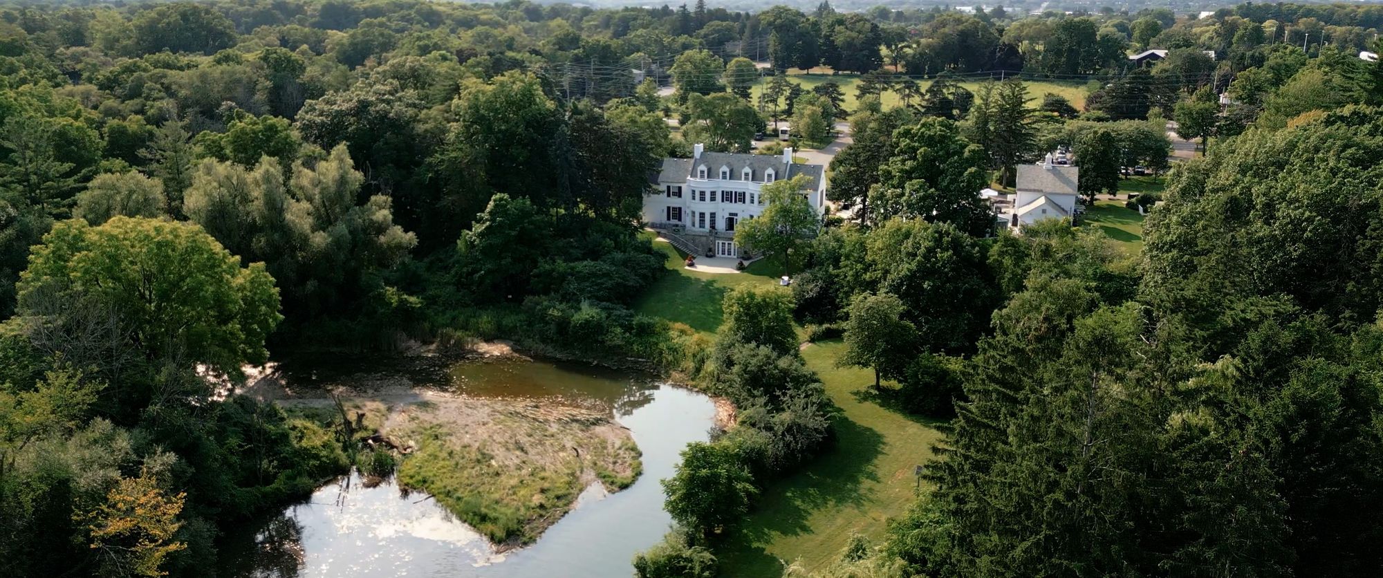 Wooded Grounds Aerial Aerial view of wooded grounds with a white building, pond, and garden paths.