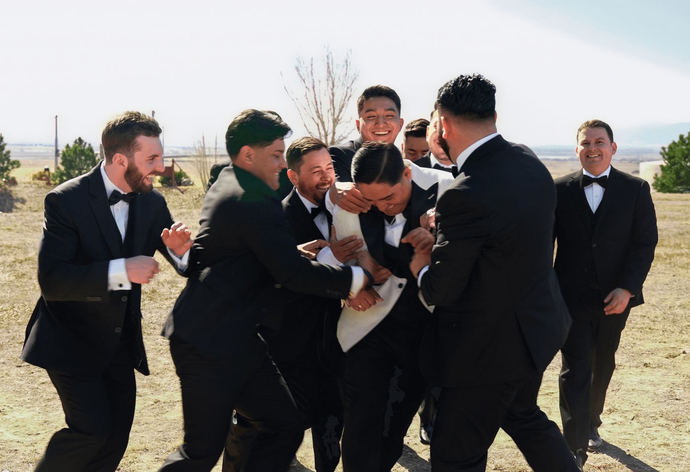 Group of men in dark suits clustered around and lifting one person in an open field.