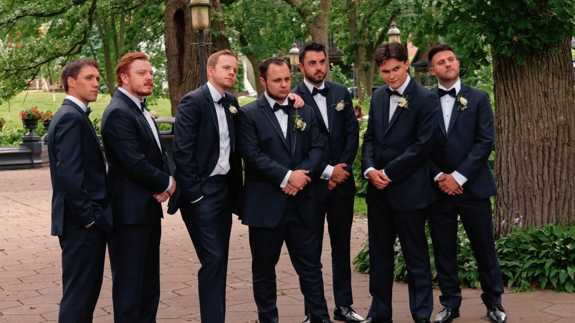 Suit Lineup Seven men in dark suits standing in a line beneath large trees.
