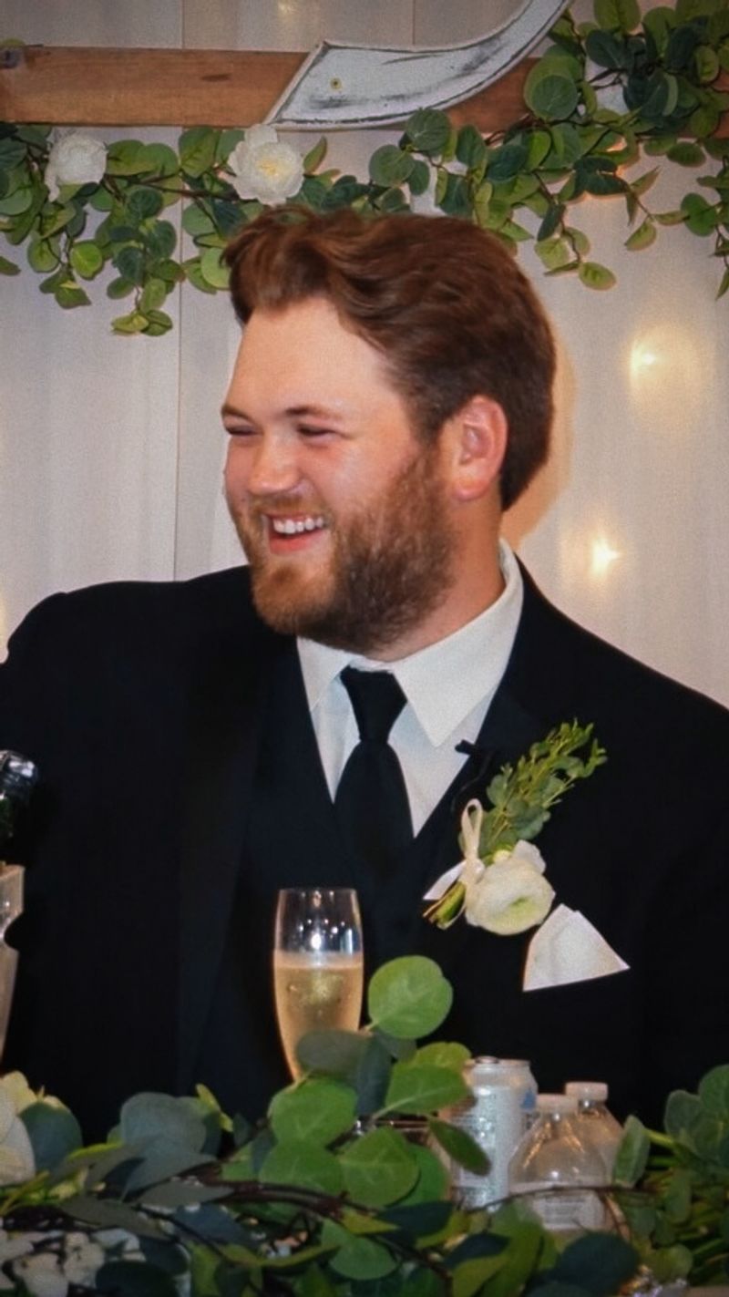 Bearded man in a dark suit seated at a decorated table with a drink and greenery.