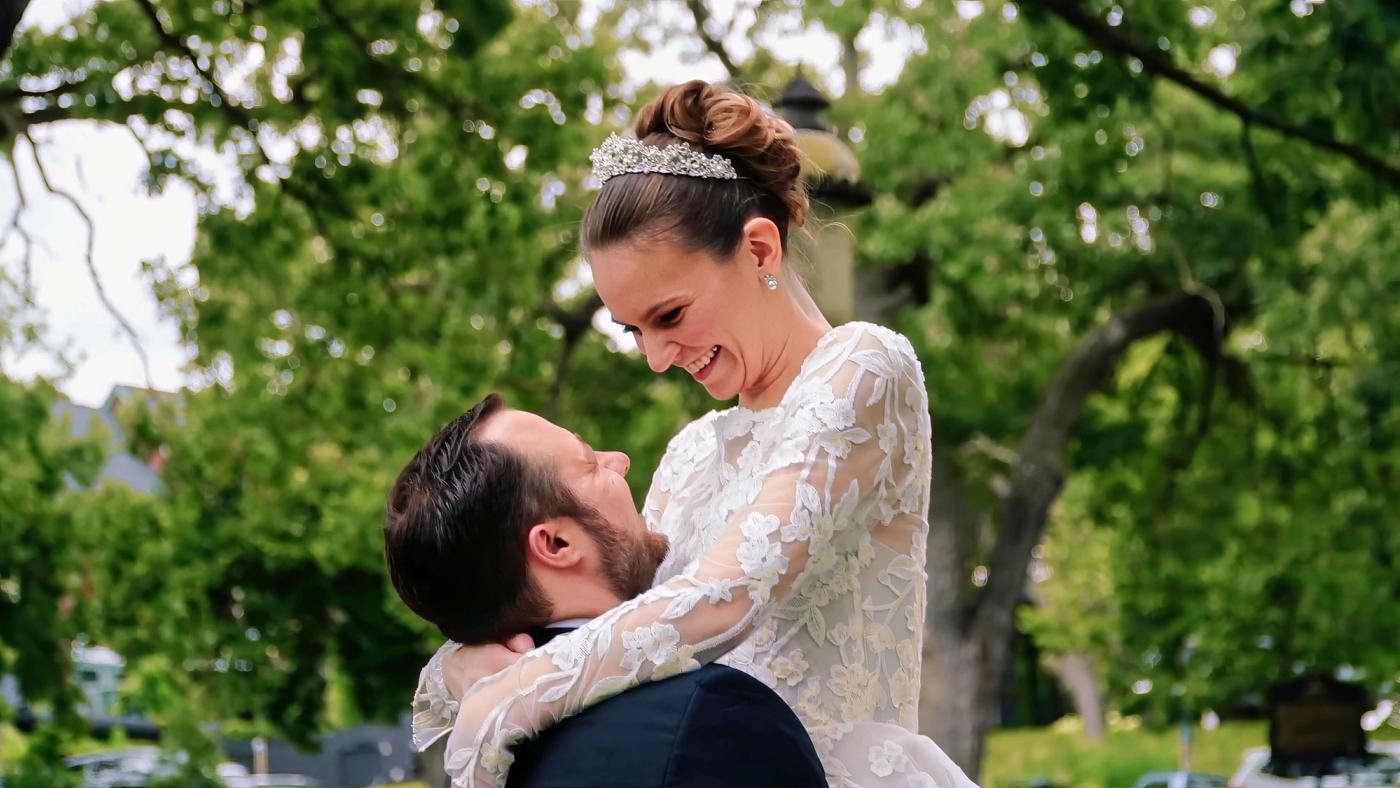 Person in a white dress held above the ground by a person in a dark suit beneath green trees.