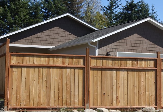 A newly constructed wooden privacy fence with dark brown posts and light cedar planks in front of a gray-brown shingled house with white trim and a dark asphalt roof, surrounded by evergreen trees under a blue sky.