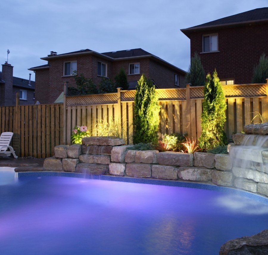 Illuminated backyard swimming pool with purple underwater lighting at dusk, featuring a natural stone retaining wall with a small waterfall, uplit evergreen trees, a wooden privacy fence with lattice top, and red brick houses in the background