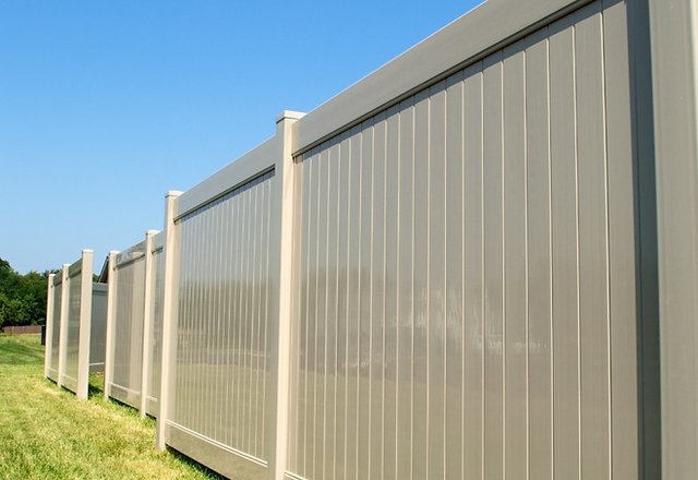 tall tan vinyl privacy fence with semi-transparent panels extending along green grass yard under clear blue sky