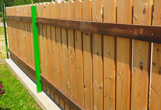 A wooden privacy fence made of vertical planks with rounded tops, reinforced by horizontal dark brown rails and a bright green fence post, shown outdoors in sunlight.