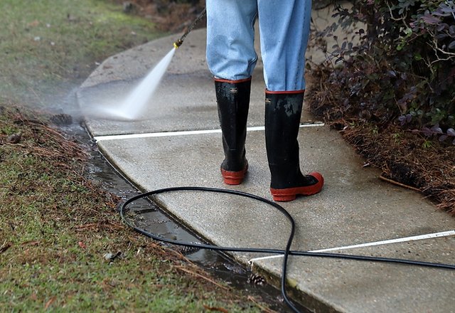person in black and red rubber boots pressure washing a concrete driveway or sidewalk with a high pressure water spray wand and hose