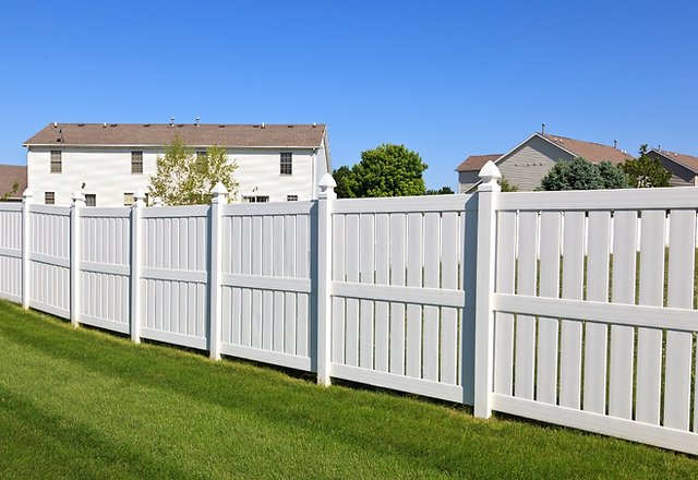 White vinyl privacy fence with decorative post caps stretching across a green lawn in a suburban neighborhood, two-story houses and trees visible under a clear blue sky.