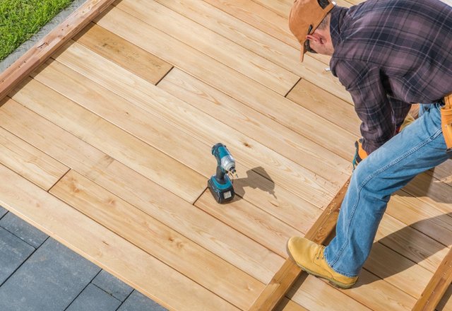 contractor installing natural cedar wood deck boards outdoors using a cordless drill while wearing safety glasses and work boots during backyard construction project