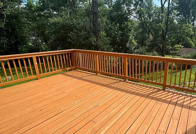 freshly stained wooden backyard deck with railings surrounded by lush green trees on a sunny day