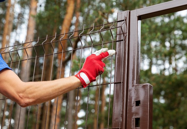 A person wearing a red work glove adjusts or secures a wire mesh panel onto a brown metal fence post outdoors, with trees visible in the background.