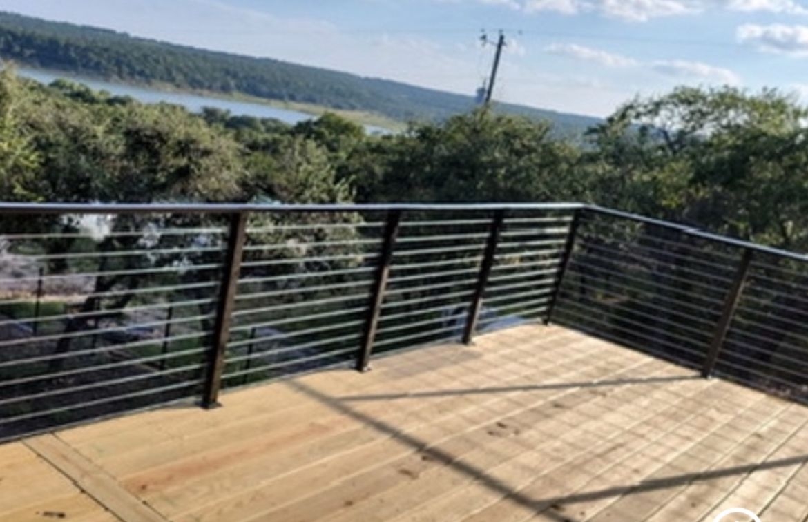 Outdoor cable railing deck with natural wood planking, black metal post railing system, and view of lake in the distance, mature trees
