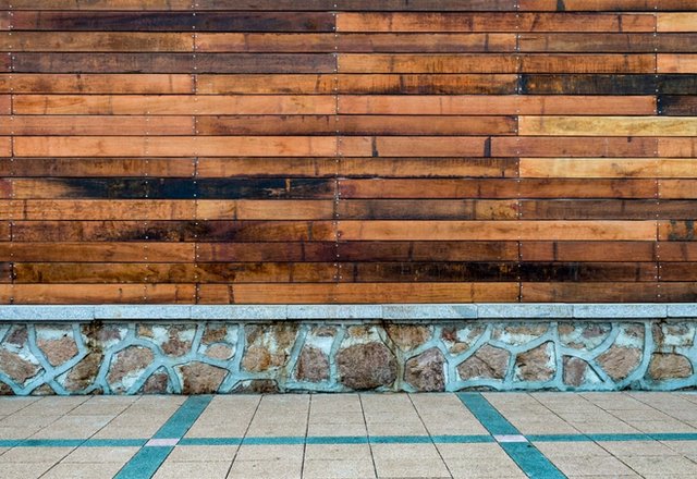 A wooden wall made of horizontal planks in varying shades of brown, mounted above a stone base, with a tiled walkway in the foreground.