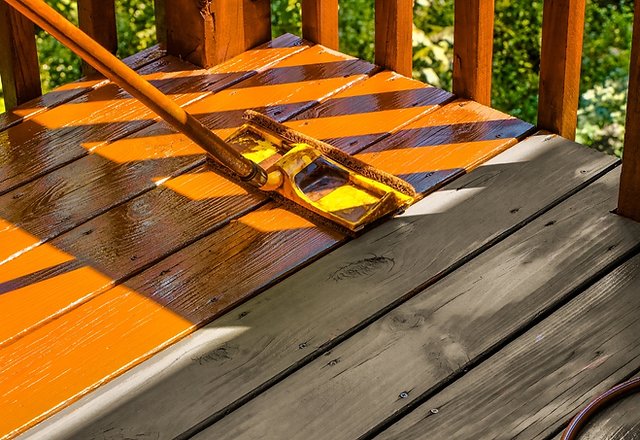 A wide staining brush spreads bright orange stain across wooden deck boards, with unstained gray planks beside it and sunlight casting shadows from the railing posts.