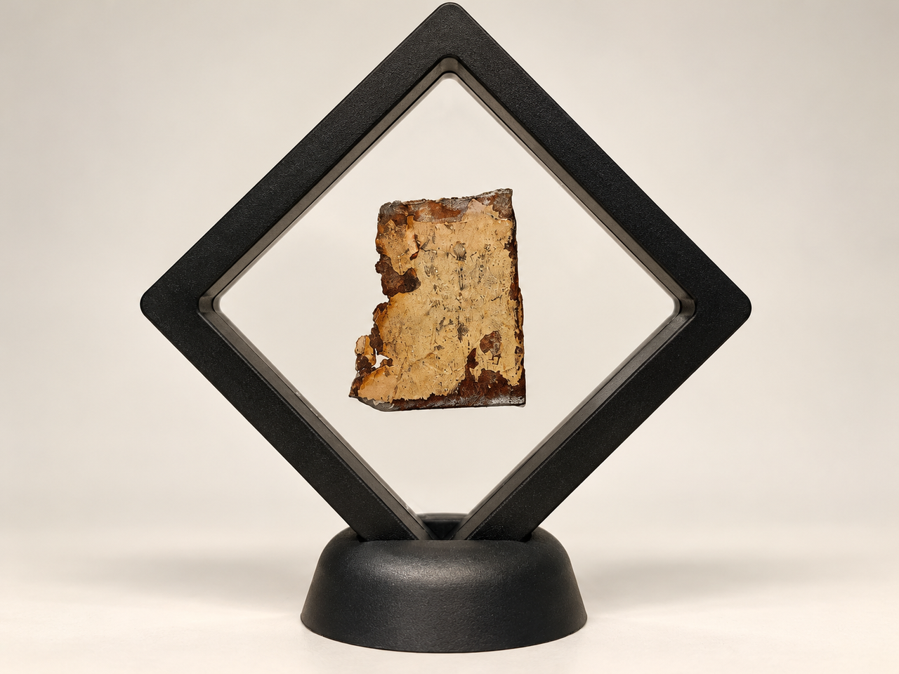 A weathered fragment of the Eiffel Tower, featuring aged beige paint and rusted edges, suspended inside a black diamond-shaped display frame against a clean, neutral background.